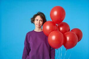 unzufrieden jung braunäugig lockig Brünette Dame Sein im Schlecht Stimmung während posieren Über Blau Hintergrund mit Luft Luftballons, verzog das Gesicht Gesicht und Verdrehen Mund foto