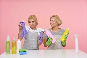Innen- Schuss von ziemlich jung blond Hausfrauen gekleidet im Basic T-Shirts und Schürzen Sitzung Über Rosa Hintergrund mit mehrfarbig Flaschen von Haushalt Chemikalien foto