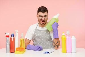 Innen- Schuss von jung dunkel behaart Mann mit kurz Haarschnitt tragen Uniform während Sitzung Über Rosa Hintergrund, vorbereiten zum Frühling Reinigung und erziehen Hand mit Haushalt Chemikalien foto