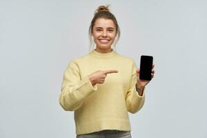 Porträt von attraktiv Mädchen mit blond Haar versammelt im Brötchen. tragen Gelb Sweatshirt und Punkte zu das Telefon Bildschirm, Kopieren Raum. breit lächelnd. Aufpassen beim das Kamera, isoliert Über Weiß Hintergrund foto