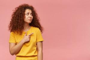 Teenager Mädchen, traurig suchen Frau mit Ingwer lockig Haar. tragen Gelb T-Shirt. Menschen und Emotion Konzept. Aufpassen und zeigen zu das richtig beim Kopieren Raum, isoliert Über Pastell- Rosa Hintergrund foto