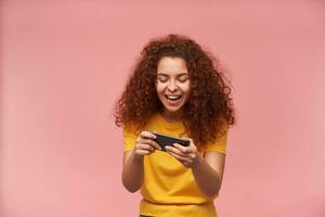Teenager Mädchen, glücklich suchen Frau mit Ingwer lockig Haar. tragen Gelb T-Shirt. spielen Video Spiel auf ihr Smartphone. haben Spaß mit Gerät. Stand isoliert Über Pastell- Rosa Hintergrund foto