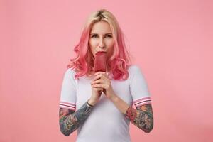 Porträt von süß Mädchen mit Rosa Haar und tätowiert Hände, suchen beim das Kamera und genießen köstlich Eis Creme, Stehen Über Rosa Hintergrund, tragen ein Weiß T-Shirt. foto