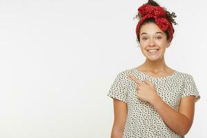 Porträt von glücklich weiblich Modell- zeigt an mit Vordergrund Finger beim leer Weiß Kopieren Raum, zeigt an Platz zum Ihre Werbung oder Werbung Text. schön Mädchen gekleidet t Hemd mit schwarz Polka Punkte foto