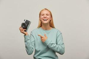 emotional Mädchen Fotograf Lachen, hält ein retro Jahrgang Foto Kamera im Hand, zeigen auf es mit Finger, bekommt Vergnügen von das Schießen Verfahren, gekleidet beiläufig Sweatshirt, Über grau Hintergrund