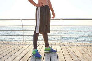 Foto von Beine von sportlich Mann, trägt ist bunt Turnschuhe und gestreift kurze Hose. Stand beim das Strand.