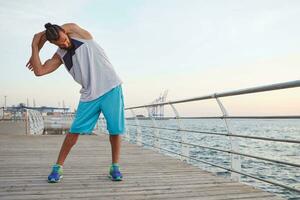 Porträt von jung sportlich bärtig Kerl tun ein sich warm laufen Vor ein Morgen Lauf beim das Strand, führt gesund aktiv Lebensstil, sieht aus weg. Fitness männlich Modell. foto