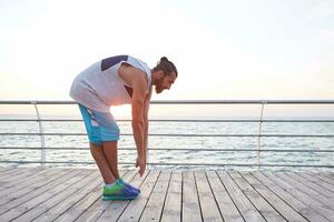 Foto von jung sportlich bärtig Kerl tun dehnen, Morgen Übungen durch das Meer, führt gesund aktiv Lebensstil. Fitness und gesund Konzept.