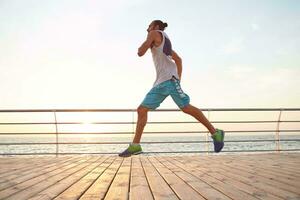 Foto von jung gut aussehend sportlich bärtig Laufen Mann , tun Morgen Übungen durch das Meer, führt gesund aktiv Lebensstil. Fitness und Sport Konzept.