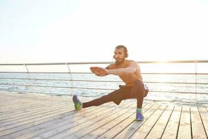 sportlich bärtig Kerl tun dehnen, Morgen Übungen durch das Meer, sich warm laufen nach laufen, Hören Liebling Musik- auf Kopfhörer, führt gesund aktiv Lebensstil. Fitness und gesund Konzept. foto