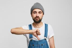 das Kerl gibt ein Negativ Bewertung, Stehen gegen Weiß Hintergrund mit ein gleichgültig nicht zufrieden Gesichts- Ausdruck, mit seine Hand zeigt an ein Daumen runter. Problem, Schlecht Stimmung foto