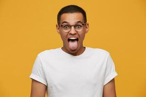 Porträt von attraktiv jung Mann im Gläser, Witze, stecken aus seine Zunge, Grimassen sieht aus lustig, trägt Weiß beiläufig T-Shirt, auf ein Gelb Hintergrund foto