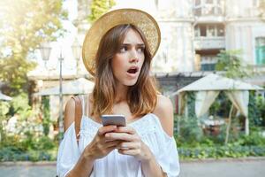 draussen Porträt von erstaunt schockiert jung Frau mit geöffnet Mund trägt stilvoll Hut und Weiß Sommer- Kleid, fühlt sich betäubt, mit Handy, Mobiltelefon Telefon und Gehen im alt Stadt foto