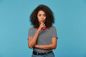 jung afro amerikanisch Frau mit afro Frisur tragen ausgezogen T-Shirt Über isoliert Blau Hintergrund fragen zu Sein ruhig mit Finger auf Lippen. Stille und Geheimnis Konzept. foto