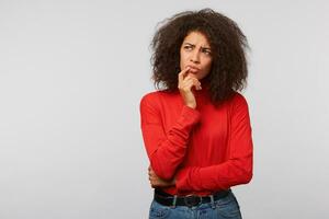 schließen oben von nachdenklich zweifelhaft jung Frau mit dunkel afro Frisur überlegt, sieht aus zu das Oberer, höher links Ecke beim leer Kopieren Raum, hält Finger in der Nähe von kinn, isoliert auf Weiß Hintergrund. foto