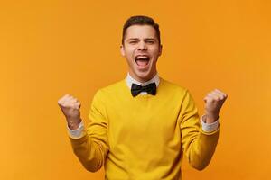 Nahansicht von emotional europäisch Mann isoliert auf Gelb Hintergrund zeigen Weiß Zähne während schreiend mit Freude und siegreich Ausdruck, halten Hände im Geste von Gewinner, suchen glücklich foto