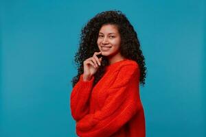 Foto von schön spanisch Latino Frau steht Hälfte Wende mit Hand berühren Gesicht sieht aus glücklich und lächelt, mit lange dunkel lockig wellig Haar im rot gestrickt Sweatshirt isoliert Über Blau Studio Hintergrund