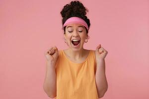 froh, überrascht Mädchen mit dunkel lockig Haar Brötchen. tragen Rosa Visier, Ohrringe und Orange Panzer oben. hat machen hoch. ballen Fäuste und schließen Augen im Aufregung. Stand isoliert Über Pastell- Rosa Hintergrund foto