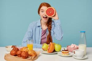 schön rothaarig Frau mit gesund Essen, Blau Hintergrund isolieren, haben Frühstück beim das Tabelle Essen Früchte Sein spielerisch Abdeckungen einer Auge mit Hälfte ein Grapefruit foto