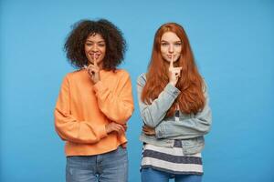 heiter jung attraktiv Frauen halten Index Finger auf ihr Lippen und lächelnd positiv beim Kamera, fragen zu behalten Geheimnis während Stehen Über Blau Hintergrund foto