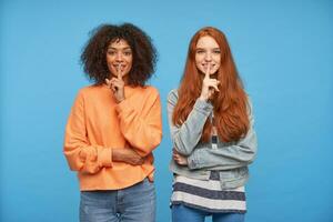 Studio Foto von positiv charmant Frauen gekleidet im beiläufig Kleider erziehen Zeigefinger zu ihr Münder und suchen fröhlich beim Kamera, isoliert Über Blau Hintergrund