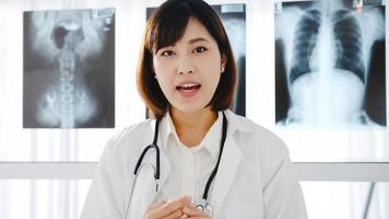 junge asiatische Ärztin in weißer medizinischer Uniform mit Stethoskop mit Computer-Laptop-Talk-Videokonferenz mit Patienten, Blick in die Kamera im Gesundheitskrankenhaus. Beratungs- und Therapiekonzept.