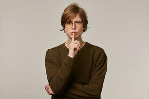 Porträt von gutaussehend, ernst männlich mit blond Haar. tragen braun Sweatshirt und Brille. zeigen Stille unterzeichnen. Menschen und Emotion Konzept. Aufpassen beim das Kamera isoliert Über grau Hintergrund foto