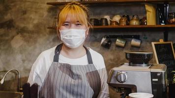 Porträt junge asiatische Kellnerin trägt eine medizinische Gesichtsmaske und fühlt sich glücklich lächeln, die nach der Sperrung im städtischen Café auf Kunden wartet. Inhaber Kleinunternehmen, Essen und Trinken, Geschäftskonzept wiedereröffnet.