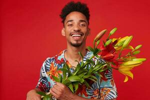 jung Lachen afrikanisch amerikanisch Mann, trägt im hawaiisch Shirt, sieht aus beim das Kamera mit glücklich Ausdruck, hält Gelb und rot Blumen Strauß , steht Über rot Hintergrund. foto