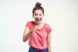 froh jung ziemlich Brünette weiblich mit beiläufig Frisur Lachen glücklich während zeigen beim Kamera mit Zeigefinger, isoliert Über Weiß Hintergrund im Rosa T-Shirt foto