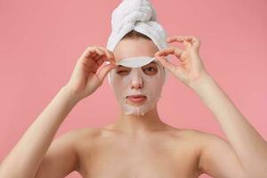 schließen oben von jung Frau mit ein Handtuch auf ihr Kopf nach Dusche, versuchen zu entfernen das Stoff Maske von das Gesicht, steht Über Rosa Hintergrund. foto