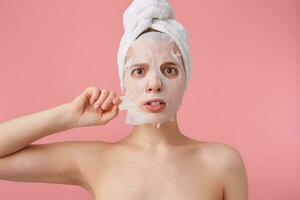 schließen oben von jung schockiert Frau mit ein Handtuch auf ihr Kopf nach Dusche, versuchen zu entfernen das Stoff Maske von das Gesicht, steht Über Rosa Hintergrund. foto