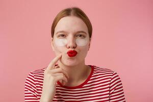 Porträt von Denken jung rothaarig Mädchen mit rot Lippen und mit Patches unter das Augen, trägt im ein rot gestreift T-Shirt, steht Über Rosa Hintergrund, Punkte zu seine Wange und lokking beim das Kamera. foto