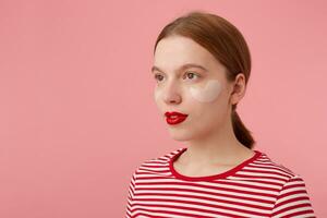 schließen oben von süß jung Ingwer Dame mit rad Lippen, trägt im ein rot gestreift T-Shirt, sieht aus beim das Kamera mit ein Ruhe Ausdruck, steht Über Rosa Hintergrund. foto