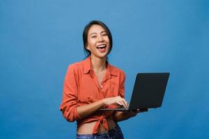 Überraschte junge Asiatin mit Laptop mit positivem Ausdruck, breites Lächeln, in Freizeitkleidung gekleidet und mit Blick auf die Kamera auf blauem Hintergrund. glückliche entzückende frohe frau freut sich über erfolg.