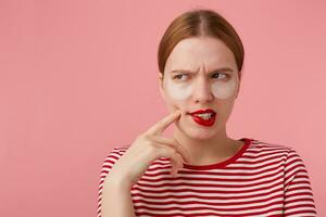 Porträt von süß Denken jung rothaarig Mädchen mit rot Lippen und mit Patches unter das Augen, trägt im ein rot gestreift T-Shirt, Wunder Was Kleid zu tragen. steht Über Rosa Hintergrund. foto