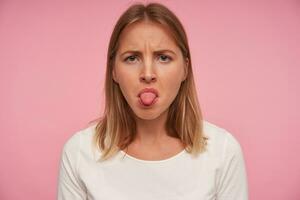 Porträt von ernst suchen ziemlich blauäugig weiblich mit blond Haar suchen beim Kamera mit unzufrieden Gesicht und zeigen Zunge, Stehen Über Rosa Hintergrund im beiläufig tragen foto