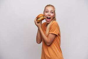 Studio Foto von jung blauäugig aufgeregt blond weiblich gekleidet im Orange T-Shirt halten groß Burger im angehoben Hände und suchen beim Kamera mit breit Augen und Mund geöffnet, isoliert Über Weiß Hintergrund