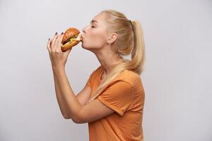 gut suchen jung blond Frau mit beiläufig Frisur halten lecker Hamburger im ihr Hände und küssen es mit geschlossen Augen, tragen Orange T-Shirt während posieren Über Weiß Hintergrund foto