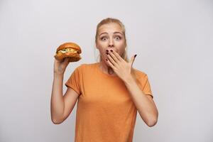 überglücklich jung ziemlich lange behaart blond Dame halten köstlich Burger im angehoben Hand und suchen beim Kamera mit breit Augen geöffnet, Abdeckung ihr Mund mit Palme Über Weiß Hintergrund foto