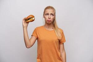 verärgert jung lange behaart blond Dame im Orange T-Shirt suchen beiseite traurig und Faltenbildung Stirn während halten ungesund Essen im angehoben Hand, Stehen gegen Weiß Hintergrund foto