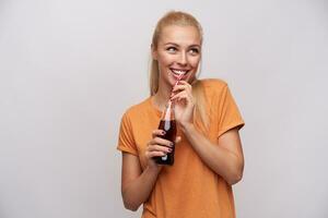 Innen- Foto von jung freudig blond weiblich mit Pferdeschwanz Frisur halten Glas Flasche von Limonade im angehoben Hände und suchen fröhlich beiseite, isoliert Über Weiß Hintergrund