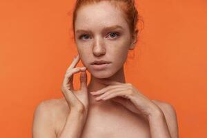Studio Foto von anmutig jung Lesekopf Dame mit foxy Haar tragen Nein bilden, erziehen Hand zu ihr Gesicht im fein Weg, berühren Wange leise während posieren Über Orange Hintergrund