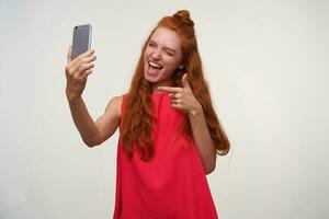 Porträt von froh jung Dame im Rosa Kleid warnen ihr wellig foxy Haar im Brötchen, halten Smartphone im angehoben Hand während Herstellung komisch Sellfie, zeigen zu Kamera mit Zeigefinger und lächelnd fröhlich foto