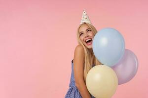 schön heiter jung lange behaart Dame im Blau Sommer- Kleid und Kegel Hut zeigen glücklich Reaktion auf bekommen genial gegenwärtig, posieren Über Rosa Hintergrund mit mehrfarbig Helium Luftballons foto