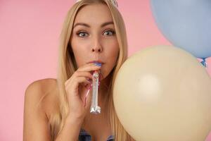 froh jung blond Frau posieren im mehrfarbig Luft Luftballons, halten Party Horn im ihr Mund und suchen beim Kamera glücklich, Gruß freunde mit Besondere Gelegenheit, isoliert Über Rosa Hintergrund foto