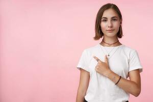 horizontal Schuss von jung ziemlich braun behaart Dame mit natürlich bilden behalten ihr Lippen gefaltet während zeigen beiseite mit Index Finger, isoliert Über Rosa Hintergrund foto