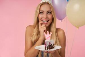 glücklich schön blond weiblich suchen beiseite mit charmant Lächeln und behalten Zeigefinger auf ihr Unterlippe, halten Stück von Kuchen während posieren Über Rosa Hintergrund mit mehrfarbig Luft Luftballons foto