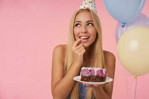 Erfreut ziemlich Frau Jubel während posieren im mehrfarbig Luft Luftballons, feiern Geburtstag mit köstlich Kuchen, suchen beiseite positiv und träumend Über Zukunft, isoliert Über Rosa Hintergrund foto