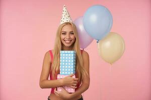 Porträt von bezaubernd positiv blond weiblich mit lange Haar halten Geschenk verpackt Kisten und zeigen ihr angenehm Emotionen, posieren Über Rosa Hintergrund im Urlaub Outfit foto